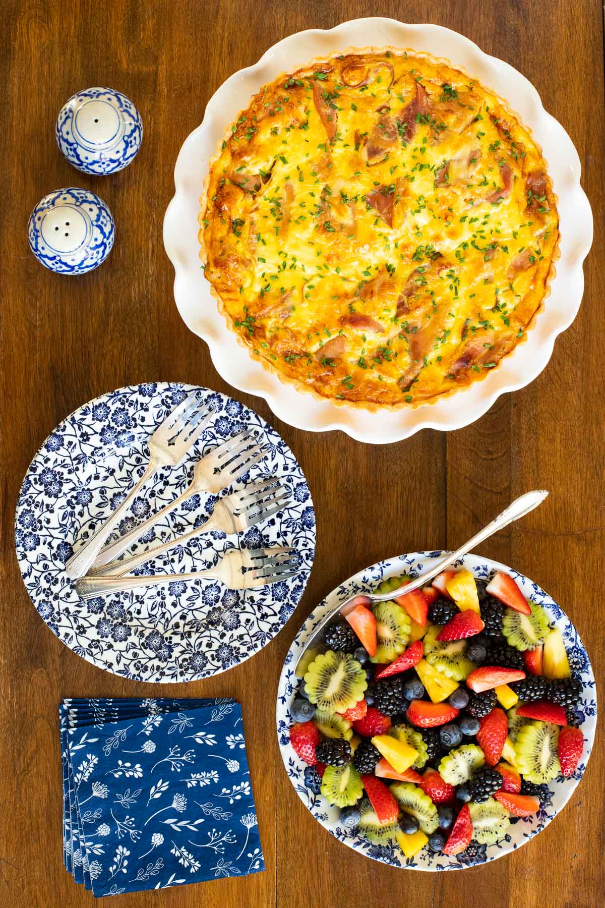 Vertical overhead photo of a Classic Quiche Lorraine with Prosciutto with a plate of fresh fruit on a wood table.