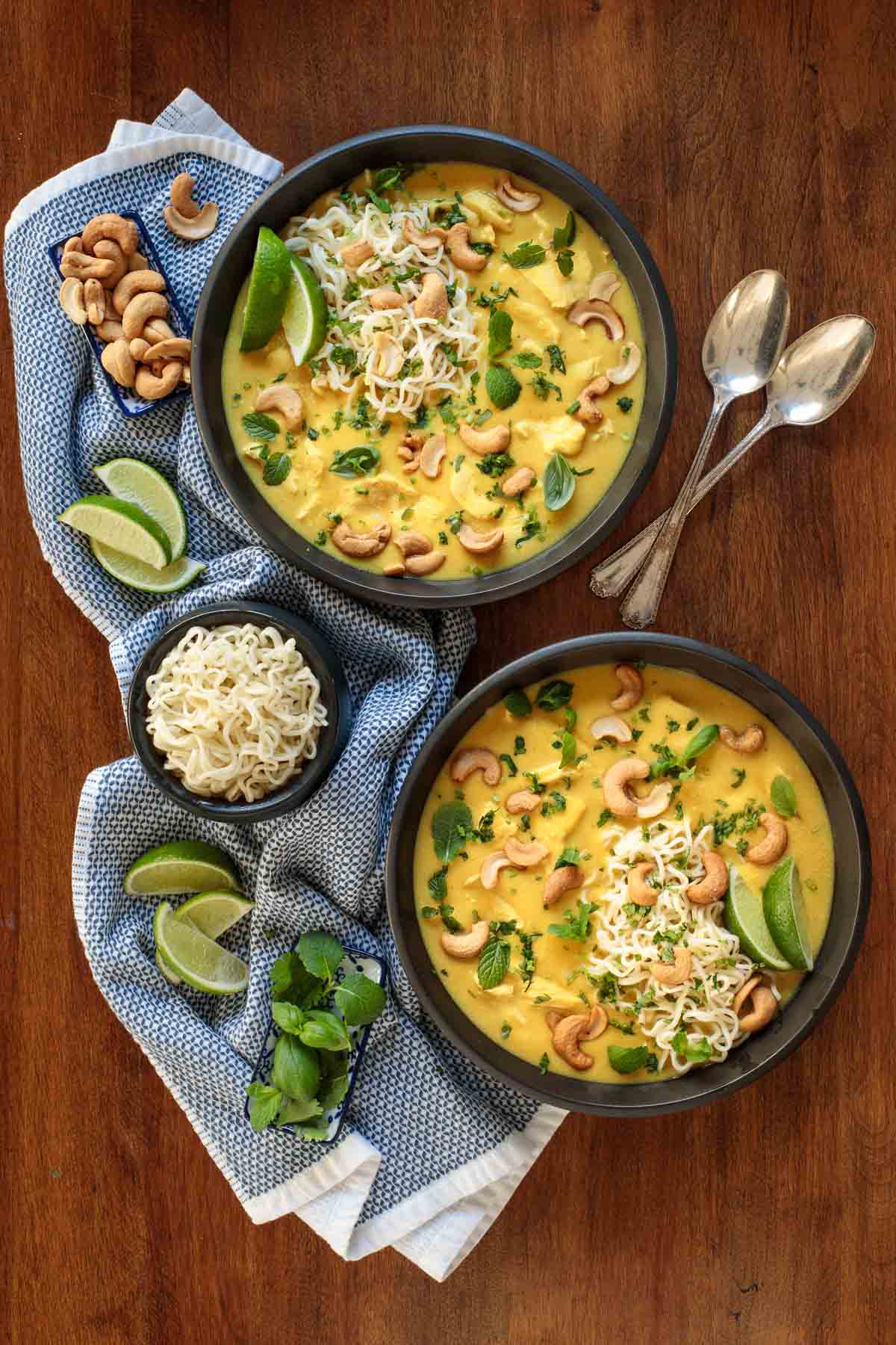 Overhead vertical photo of Coconut Curry Chicken Ramen in black serving bowls on a wooden table with garnishes of lime wedges and whole cashews.