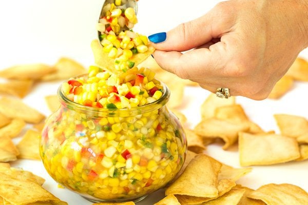 Photo of a glass jar of Trader Joe's Corn and Chile Salsa surrounded with pita chips and a hand holding a chip filled with the salsa above the jar.