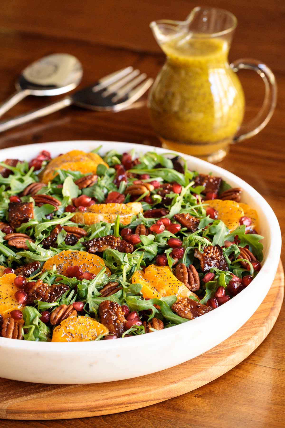 Vertical closeup photo of a Cranberry Clementine Arugula Salad in a white granite serving bowl with a small pitcher of dressing in the background.