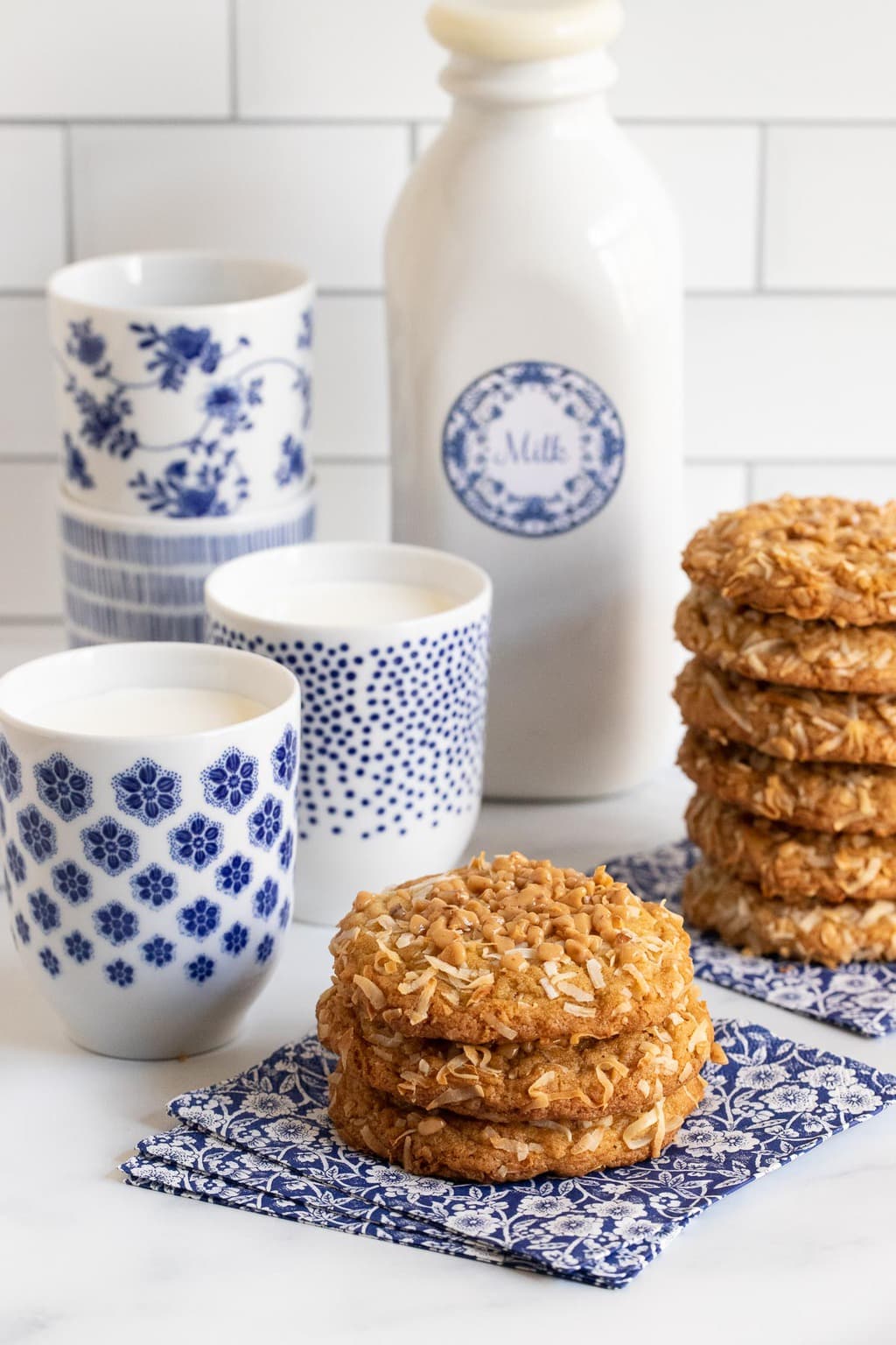 Vertical photo of stacks of Crispy Chewy Carolina Coconut Cookies on blue and white patterned napkins with cups and a bottle of milk in the background.