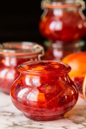 Glass jar filled with vibrant blood orange marmalade, surrounded by other jars and fresh blood oranges on a marble surface.