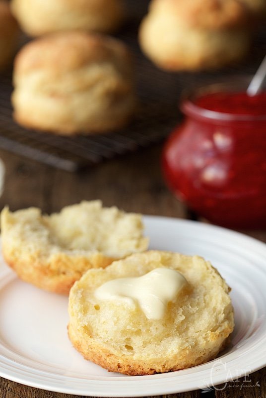 Vertical photo of a Ridiculously Easy Buttermilk Biscuit with melting butter on a white plate with jam in the background.