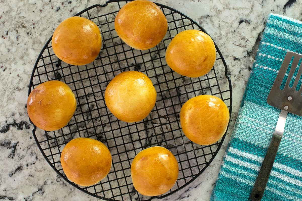 Overhead photo of a batch of Easy Buttermilk Brioche Buns on a black cooling rack sitting on a granite countertop with a turquoise patterned dish cloth and spatula.