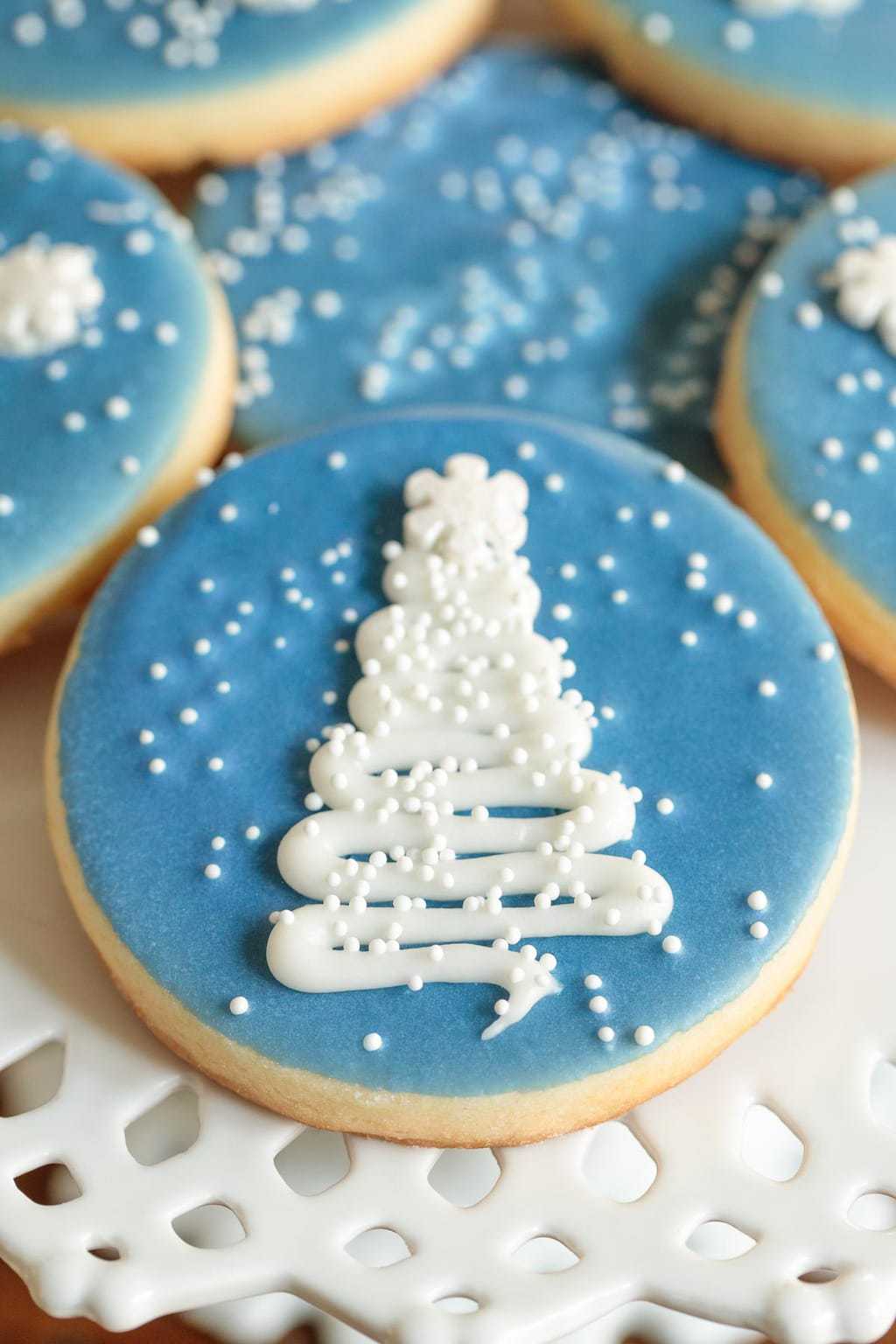 Super closeup of a white lace cake stand filled with Easy Decorated Christmas Cookies with a blue background and a white Christmas tree.