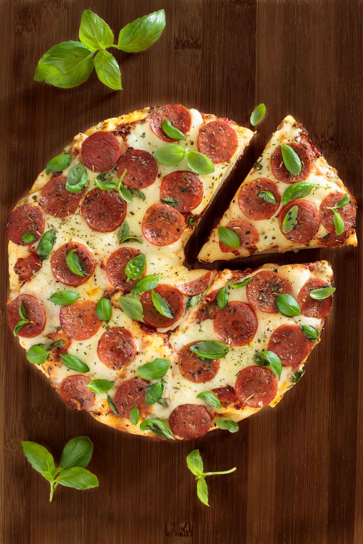 Overhead vertical photo of Easy Deep Dish Pepperoni Pizza on a wooden cutting board garnished with fresh basil leaves.