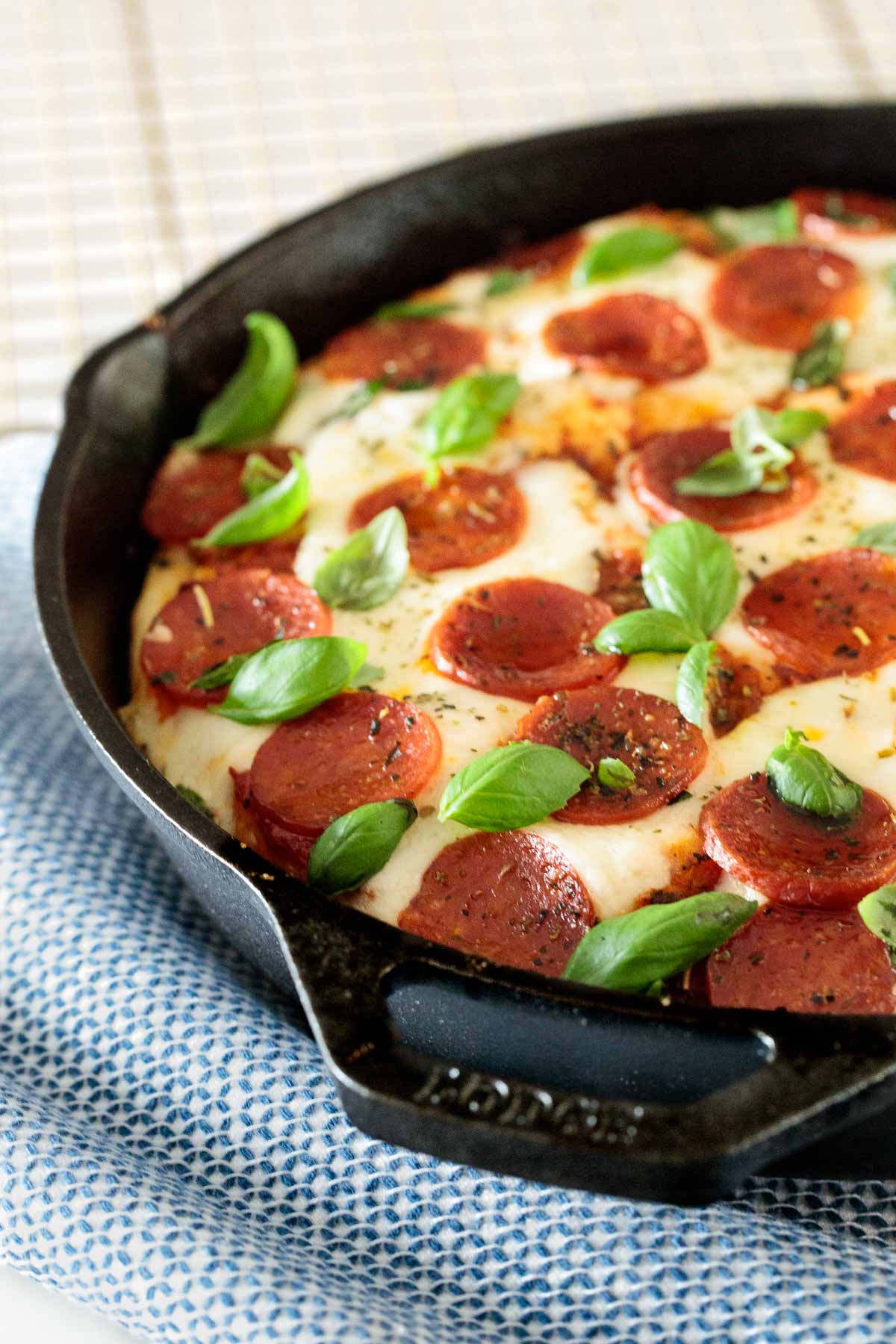 Side image of a deep dish pepperoni pizza in a cast iron pan with a blue and white dish towel underneath it.