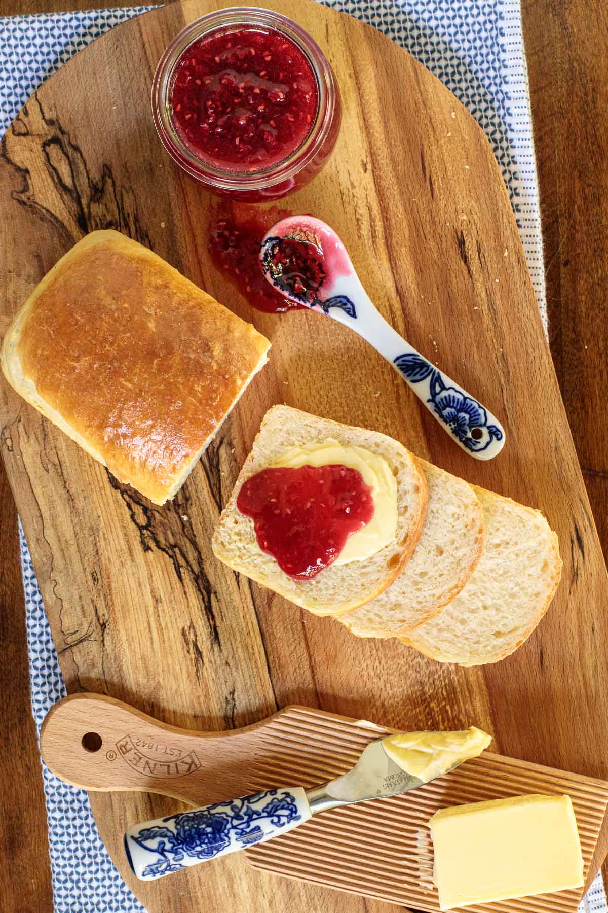 Overhead vertical photo of Honey Brioche Bread sliced on a wooden cutting board with raspberry jam and butter.