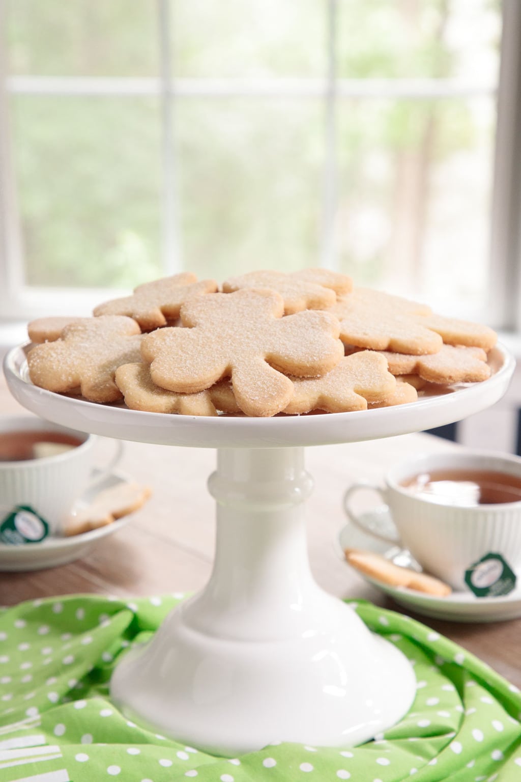 Photo of a white pedestal stand filled with Easy Irish Shortbread Cookies surrounded by a green and white cloth and cups of Irish tea.