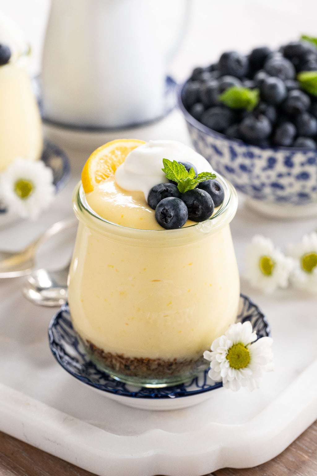 Vertical closeup photo of Easy Lemon Curd Mouse in a small glass Weck jar garnished with a lemon wedge, blueberries, mint leaves and whipped cream.