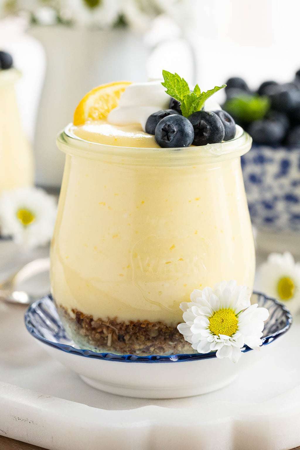 Vertical closeup photo of Easy Lemon Curd Mouse in a small glass Weck jar garnished with blueberries, mint leaves and flowers.