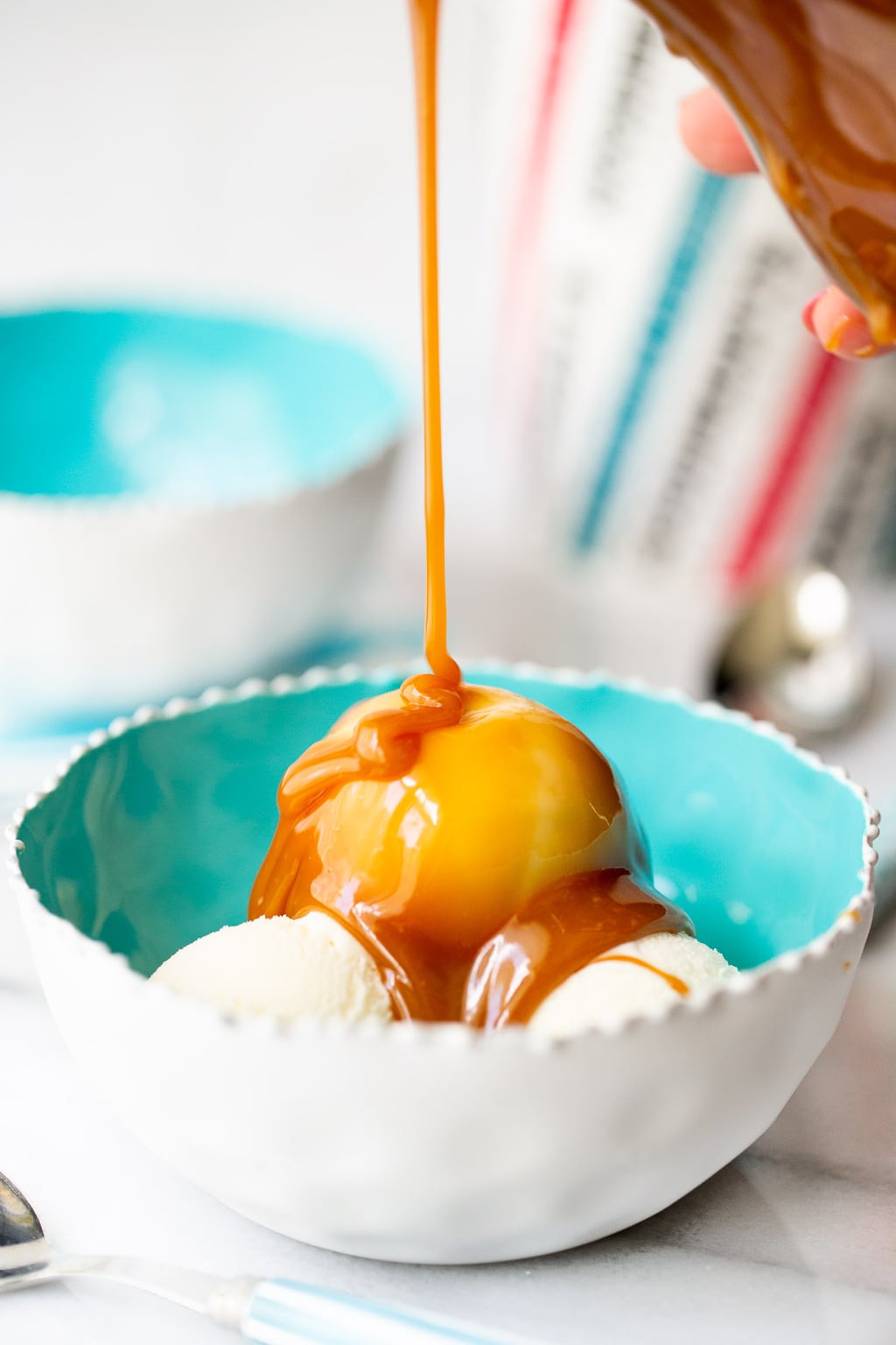Vertical closeup of Easy Microwave Caramel Sauce being poured over vanilla ice cream in a turquoise serving bowl.