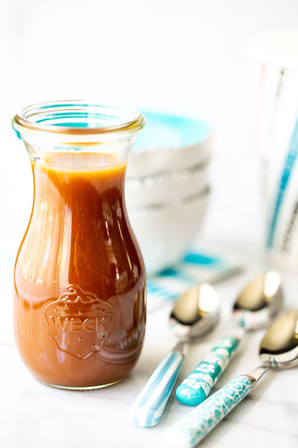 Vertical photo of a Weck glass carafe filled with Easy Microwave Caramel Sauce and surrounded by turquoise-trimmed bowls and utensils.