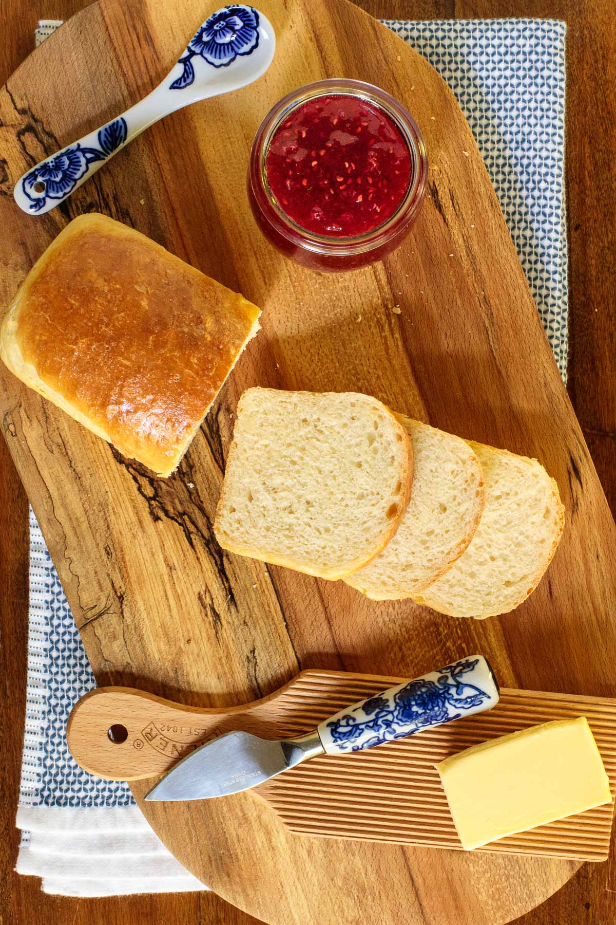 Overhead vertical photo of No-Knead Brioche Bread on a wooden cutting board with a jar of raspberry jam and Irish butter along side.