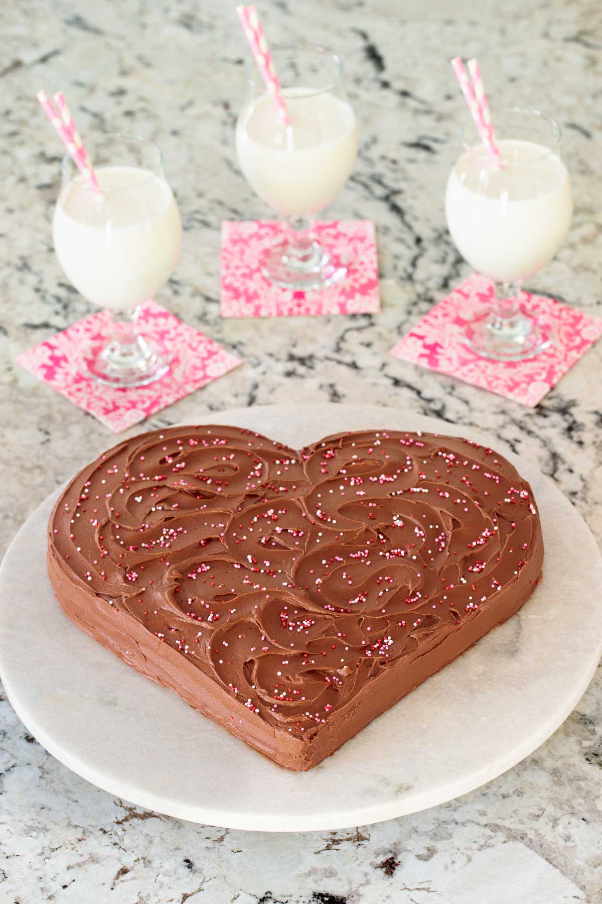 Vertical photo of a Easy One Bowl Chocolate Heart Cake on a round marble slab with glasses of milk in the background