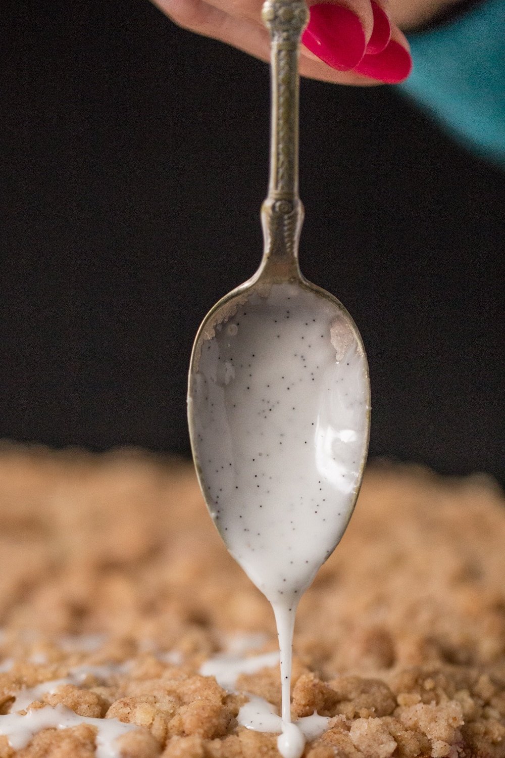 Vertical closeup photo of a spoon with Vanilla Bean Drizzle being poured over an Easy Overnight Coffee Cake.