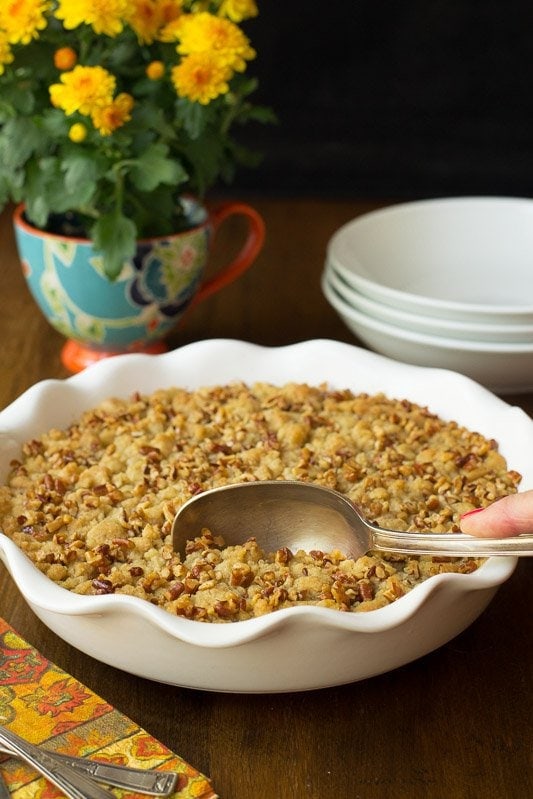 Photo of a dish of Easy Pumpkin Praline Cobbler with a hand scooping it out. Serving dishes and a mum flower is in the background.