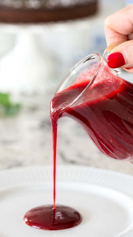 Vertical closeup photo of a glass pitcher pouring Easy Raspberry Coulis onto a white dessert plate.