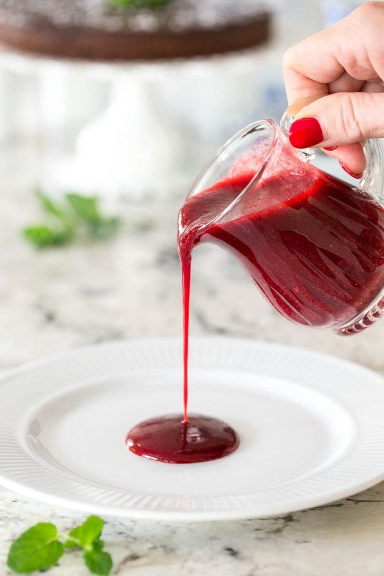 Vertical photo of Easy Raspberry Coulis in a glass pitcher poured onto a white serving plate.
