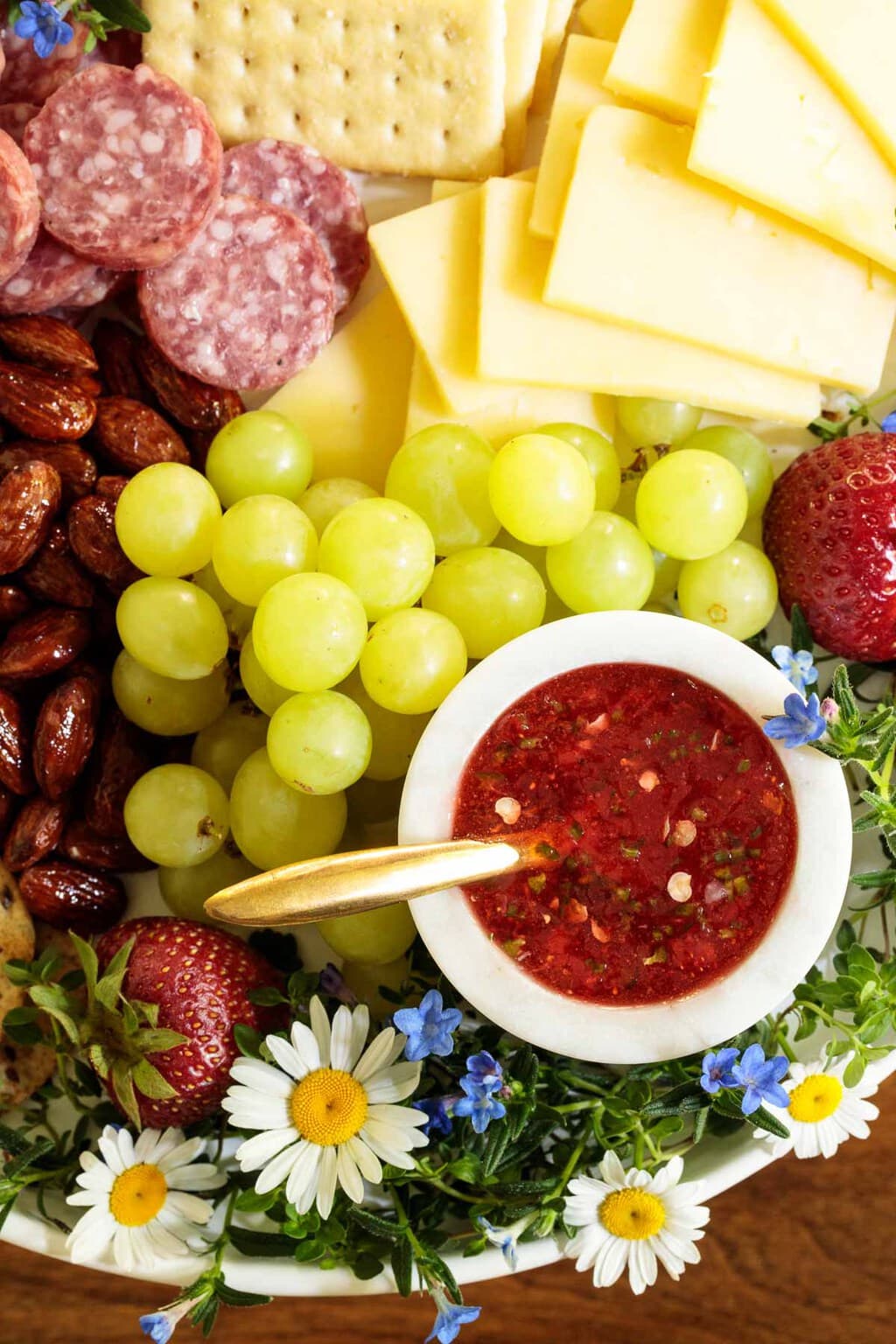 Overhead photo of an appetizer tray featuring Easy Strawberry Jalapeño Jam in a white dish surrounded by appetizer nibbles and decorative flowers.