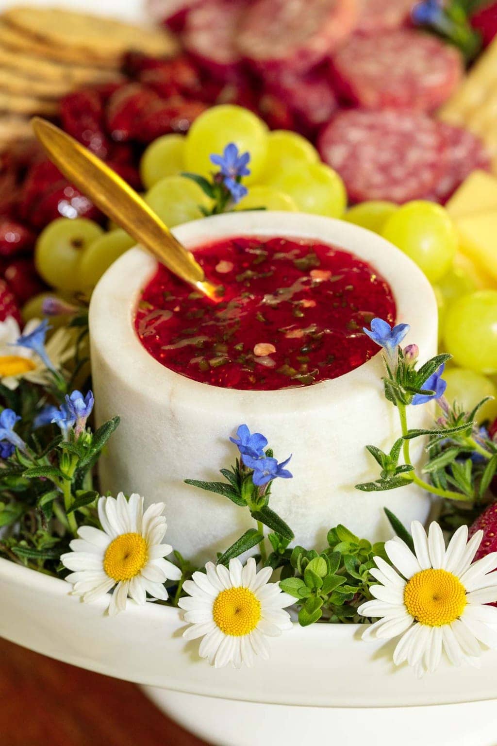 Photo of an appetizer plate featuring Easy Strawberry Jalapeño Jam surrounded by decorative flowers in the foreground and appetizer snacks in the background.