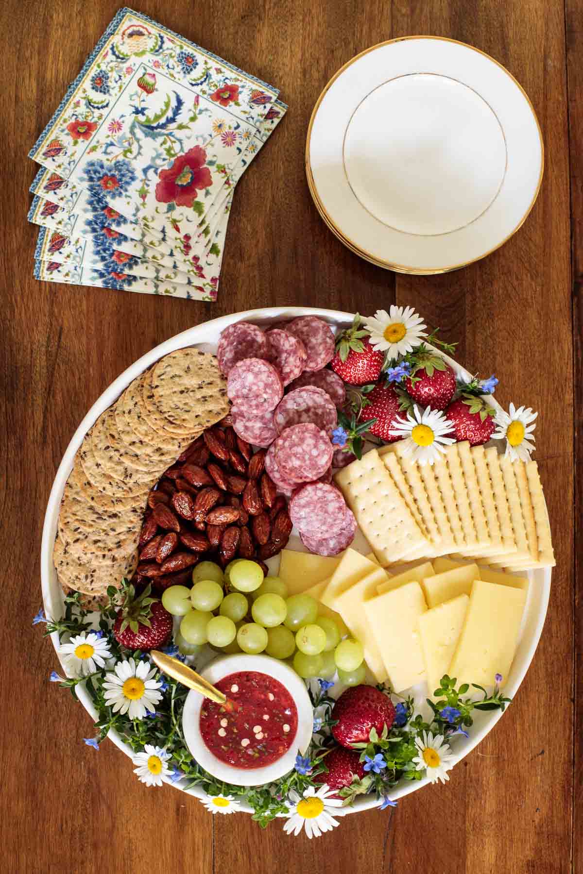 Overhead photo of an appetizer tray featuring Easy Strawberry Jalapeño Jam surrounded by appetizer snacks and decorative flowers on a wood table.