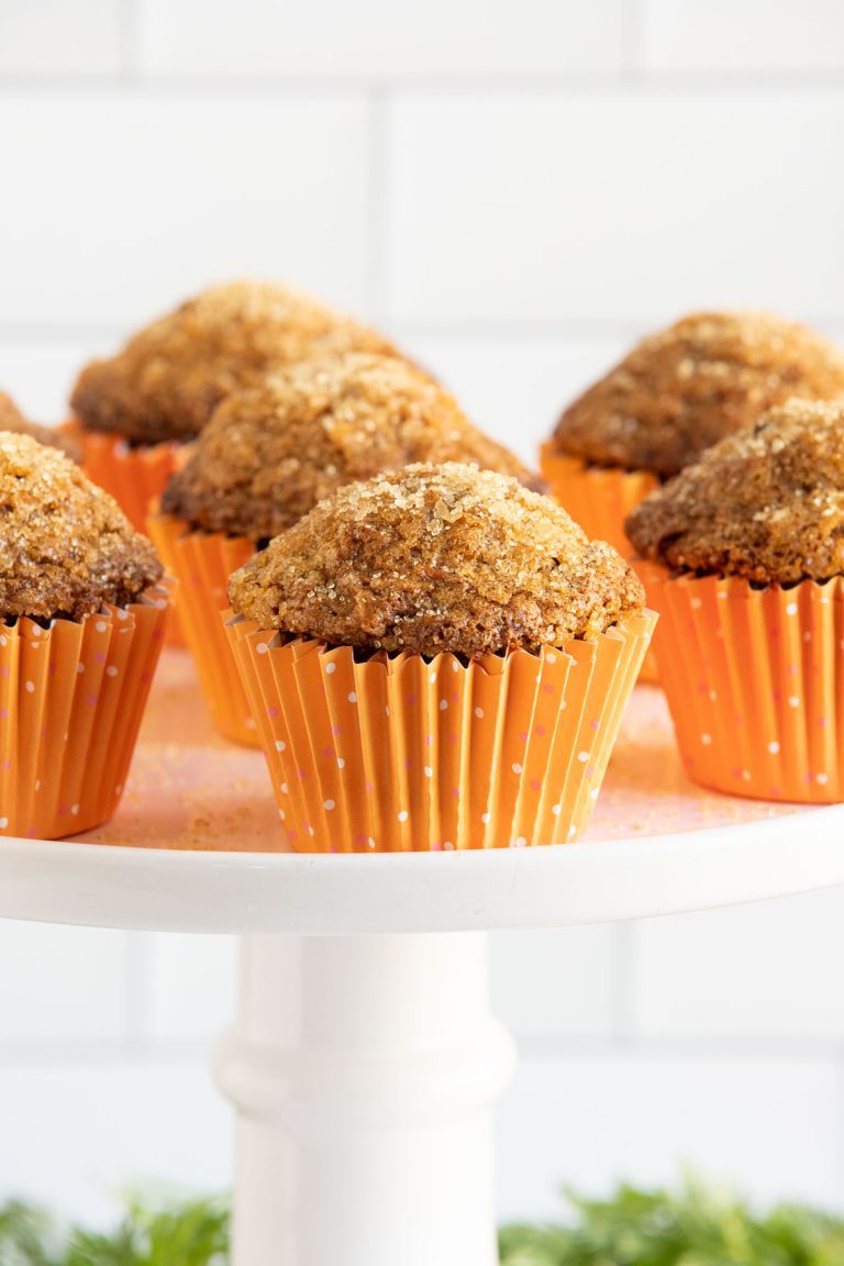 Vertical closeup photo of a batch of Sugar Top Carrot Muffins in orange cupcake liners on a white pedestal display platter.