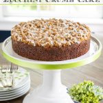 Vertical photo of a Easy Zucchini Crumb Cake on a white pedestal cake stand trimmed in pale green ribbon.