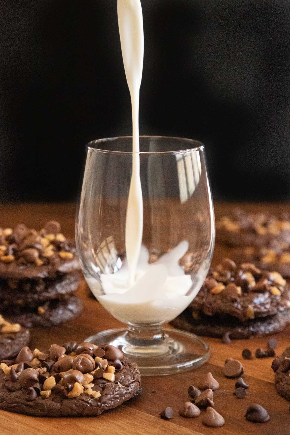 Vertical photo of milk being poured into a glass surrounded by Flourless Toffee Chocolate Cookies on a wood platter.