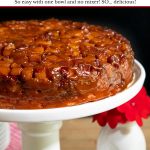 Vertical closeup photo of a French Caramel Apple Cake on a white pedestal cake stand against a black background.