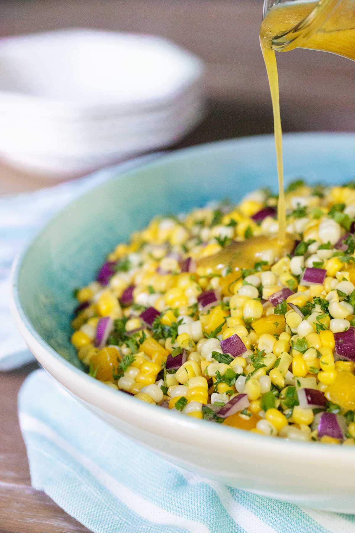 Close up photo of a white and turquoise serving bowl filled with Fresh Corn Salad. Salad Dressing is being poured over the salad.