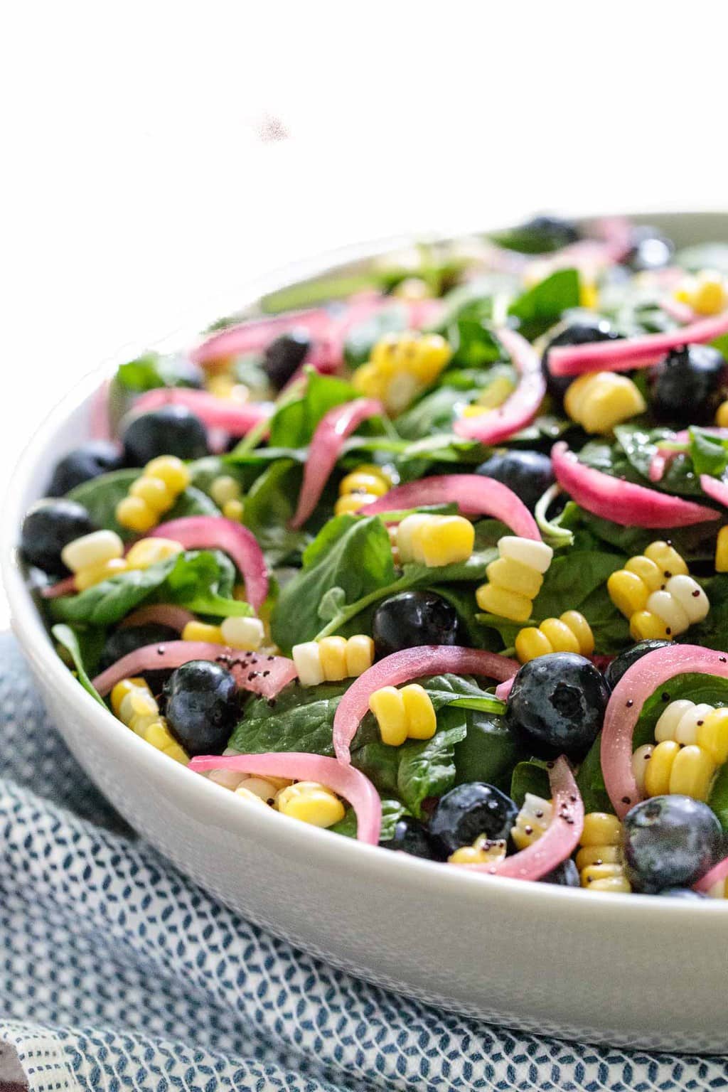 Vertical photo of Fresh Corn and Blueberry Spinach Salad in a white serving bowl on a blue and white patterned towel.