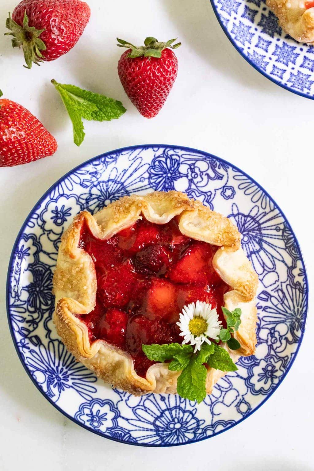 Overhead vertical photo of a Fresh Strawberry Galette on a blue patterned plate surrounded by fresh strawberries.