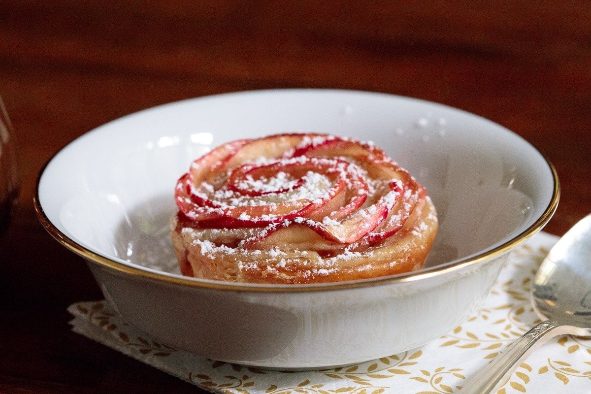 Photo of a Glazed Puff Pastry Apple Rose in a white serving bowl dusted with powdered sugar.