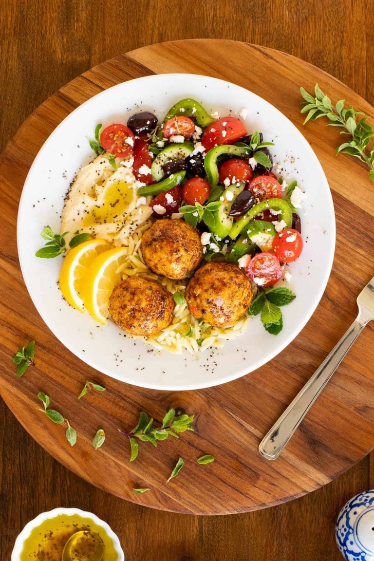 Overhead vertical photo of a serving bowl with Greek Chicken Meatballs on a bed of orzo with a Greek veggie salad.