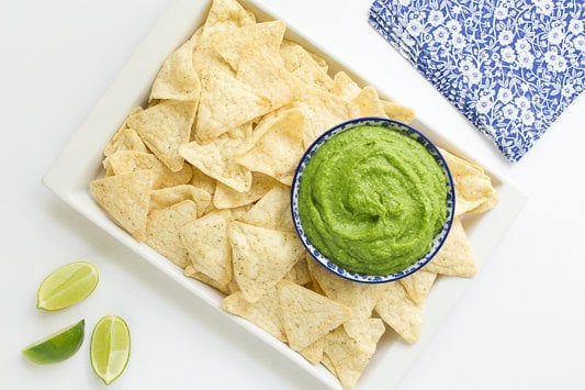 Horizontal photo of a rectangular platter of chips with a bowl of Guasacaca (Venezuelan Guacamole) in the right side of the platter. Limes and napkins surround the platter.
