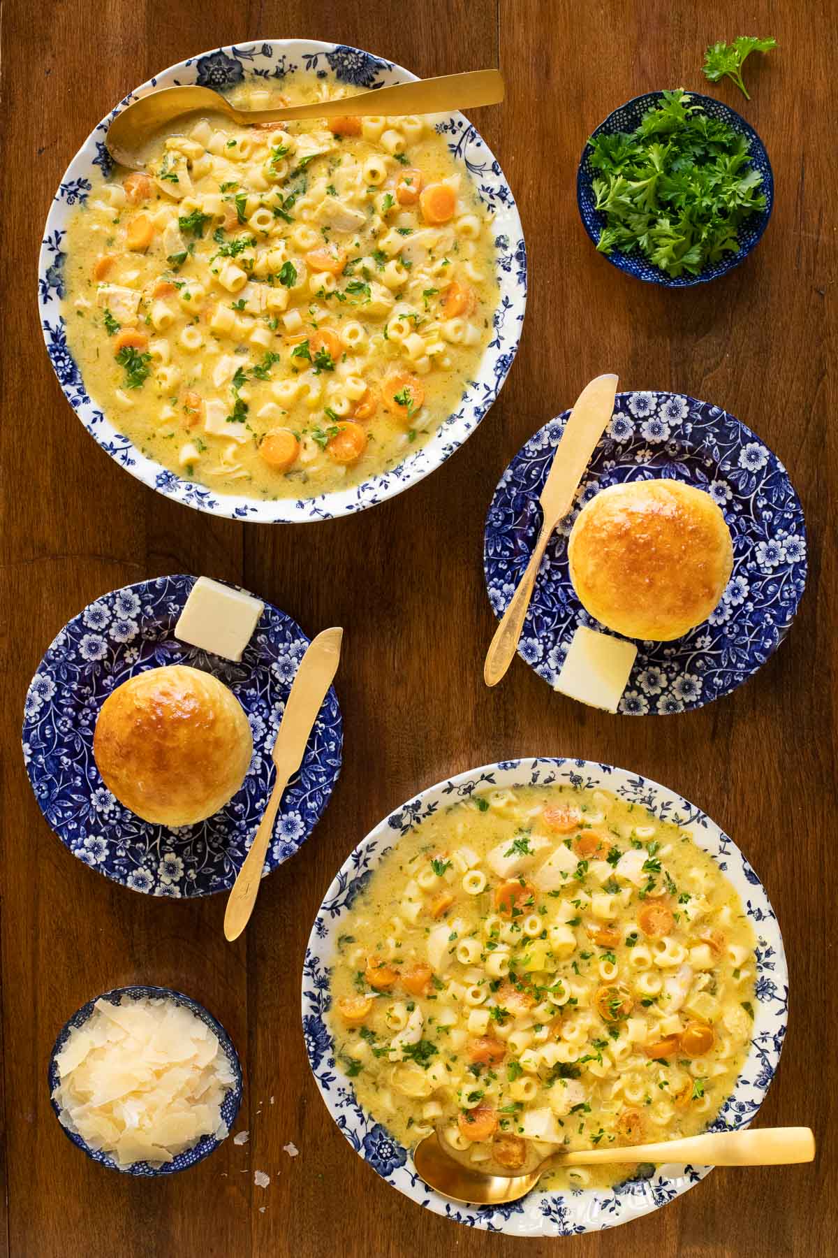 Overhead vertical photo of Creamy Chicken Pasta Parmesan Soup in blue and white serving bowls with fresh rolls and garnishes on a wood table.