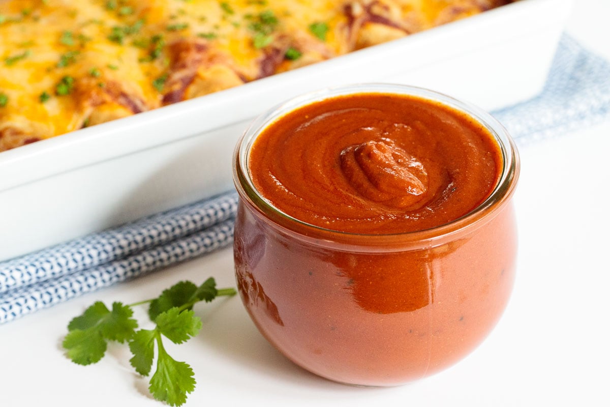 Photo of a glass jar of 10-Minute Homemade Enchilada Sauce with a white dish of enchiladas in the background and a sprig of cilantro in the foreground.
