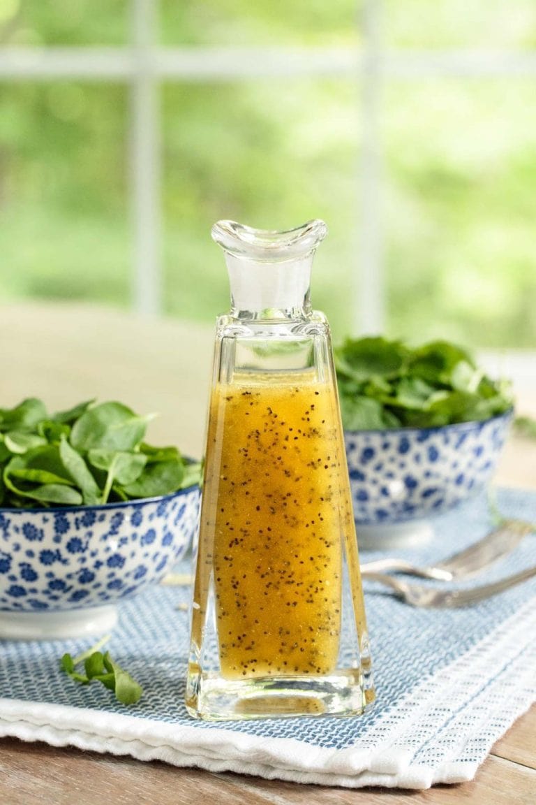 Vertical picture of Honey Cider Poppy Seed Dressing in a glass jar with bowls of fresh spinach