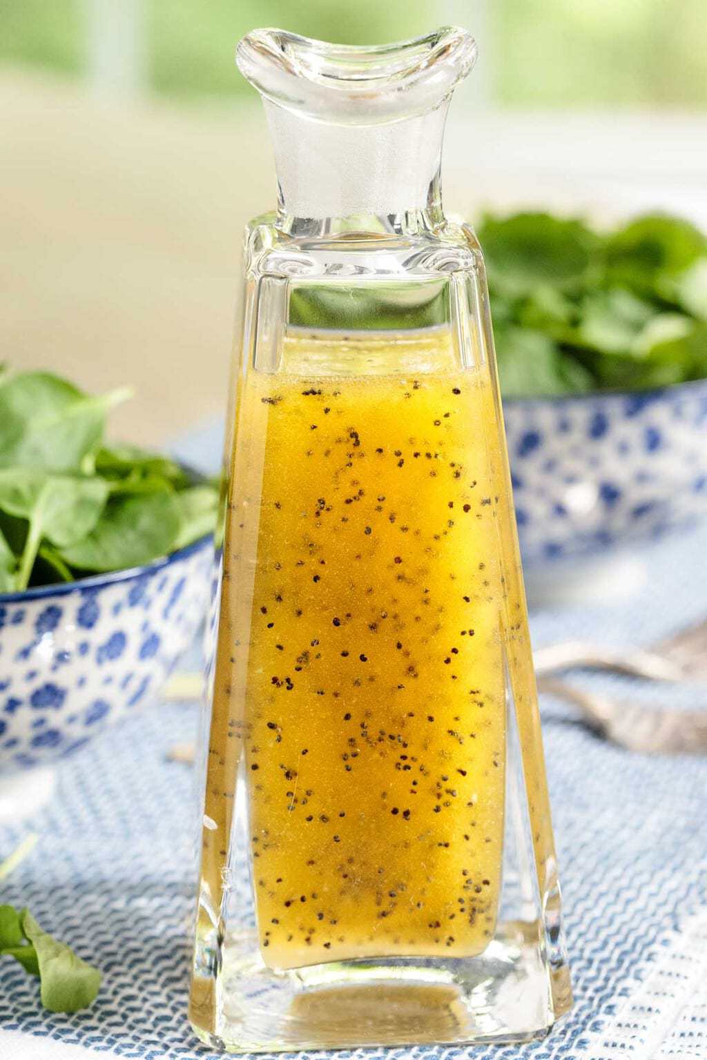 Photo of a glass decanter of Honey-Cider Poppy Seed Dressing with two spinach leaf salads in the background.