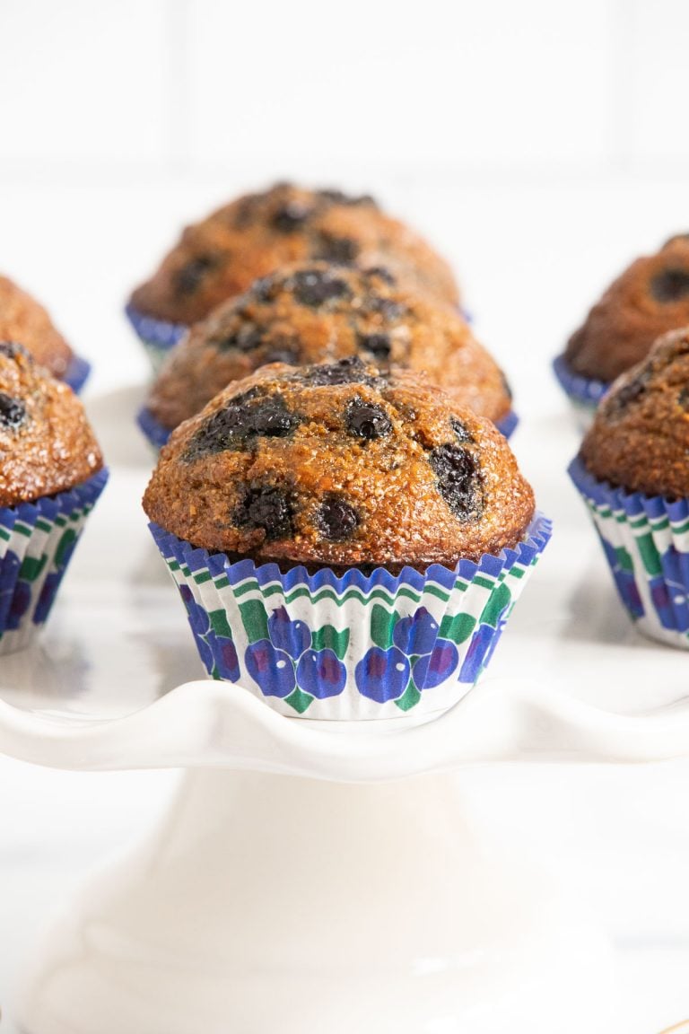 Vertical photo of a batch of Honey-Glazed Blueberry Bran Muffins on a white pedestal plate.