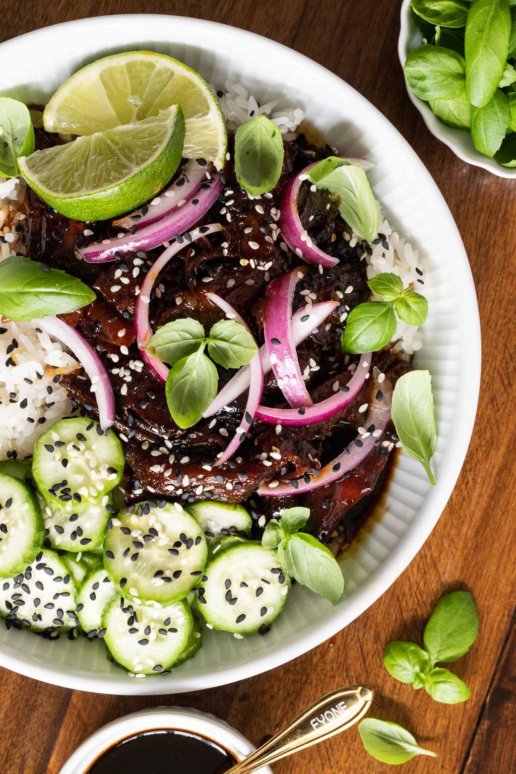 Overhead vertical photo of a bowl of Honey Soy Braised Pork with sliced cucumbers, lime wedges, pickled red onions and fresh basil leaves