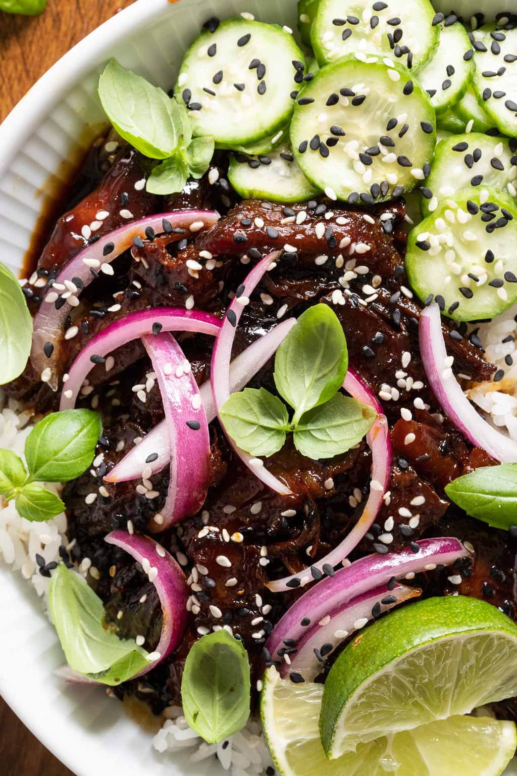 Overhead vertical closeup photo of Honey Soy Braised Pork garnished with lime wedges, pickled red onions, sesame seeds and mini cucumbers.