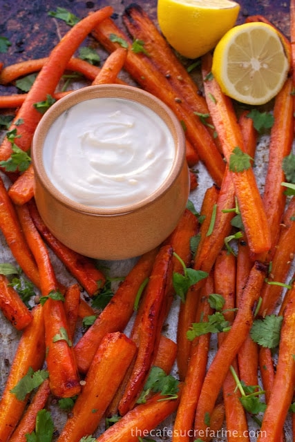 A closeup photo of a pan of Honey Roasted Carrots with a dish of lemon tahini dressing in the center and lemon halves as a garnish.