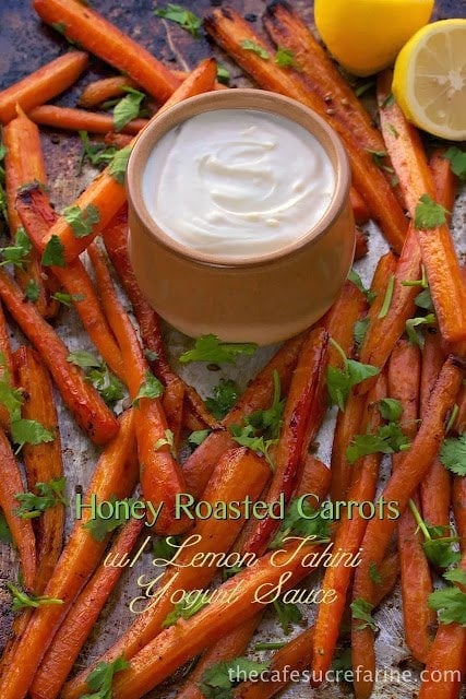 A vertical photo of a baking pan filled with Honey Roasted Carrots with a small jar of lemon tahini yogurt sauce in the middle and a graphic title at the bottom of the photo.