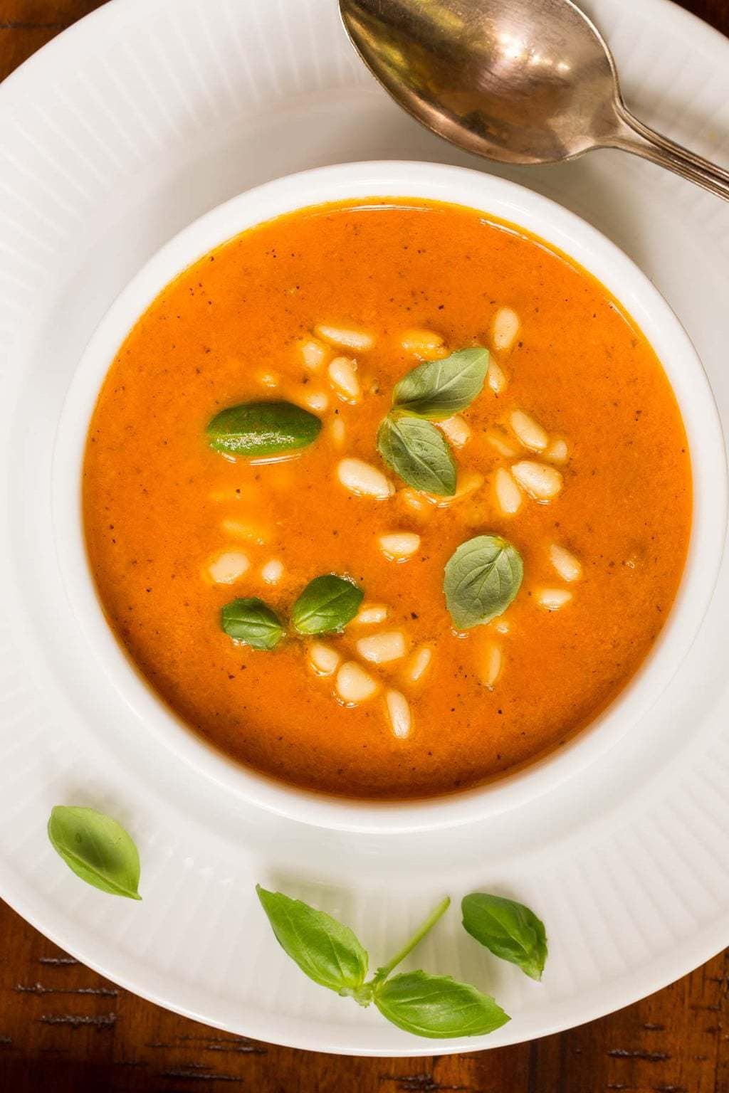 Overhead closeup photo of a bowl of Italian Sun Dried Tomato Soup in a white serving dish and plate. The plate and soup are garnished with fresh basil leaves.