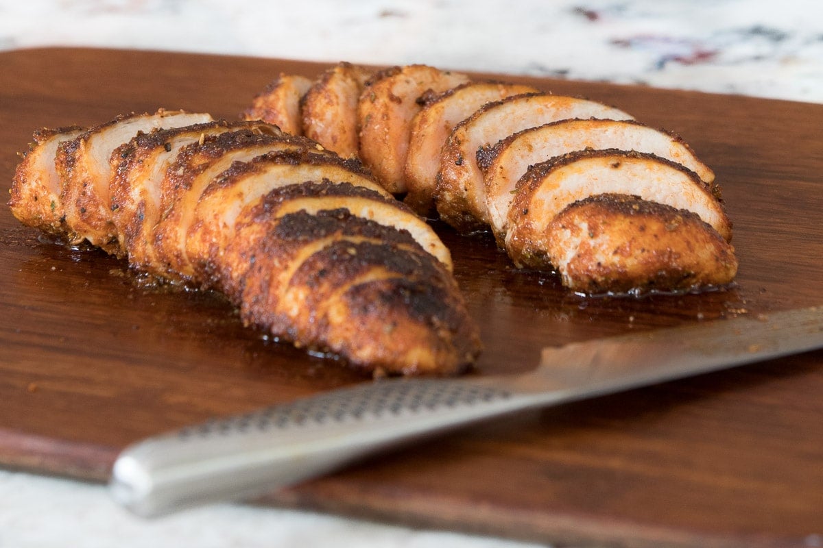Horizontal photo of sliced Juicy Tender Restaurant Style Chicken Breasts on a dark wood cutting board with a Global knife in the foreground.
