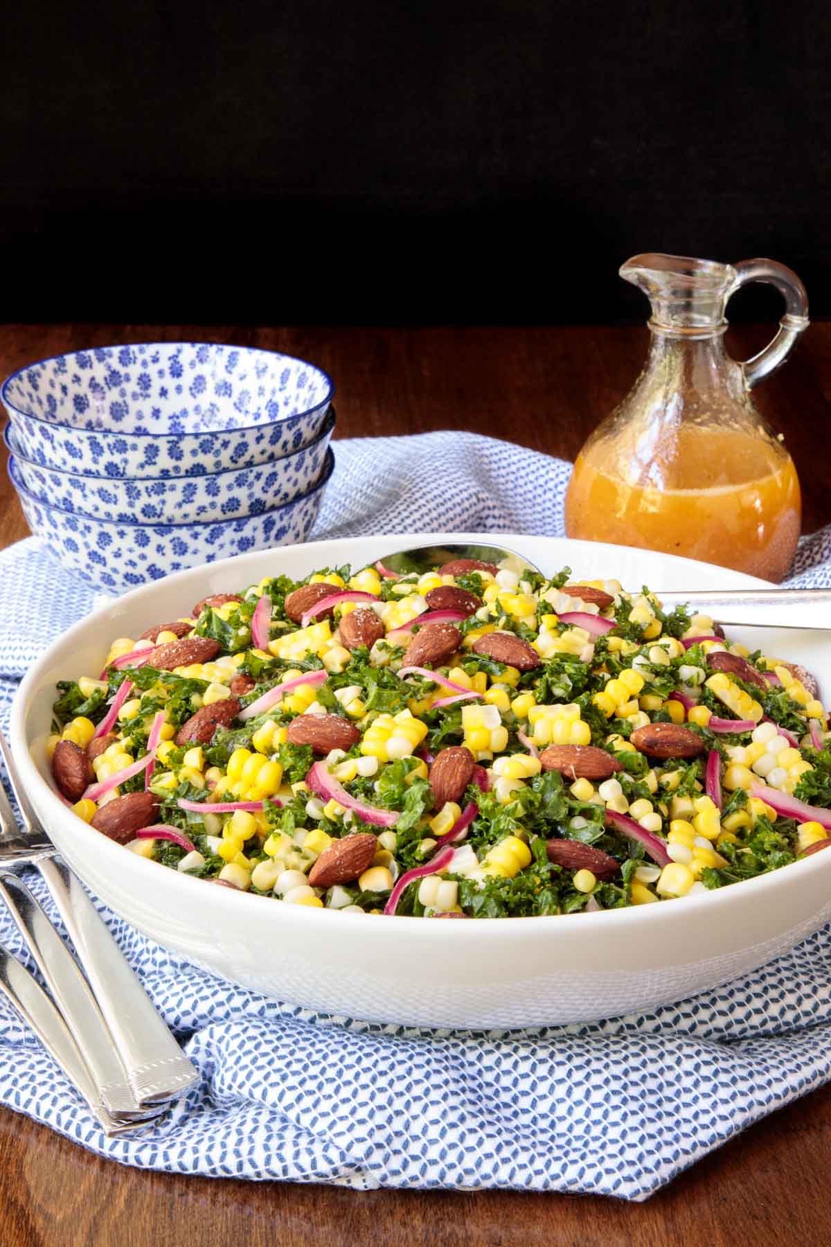 Photo of a Kale, Corn and Toasted Almonds Salad on a blue and white patterned napkin with dressing and serving bowls in the background.