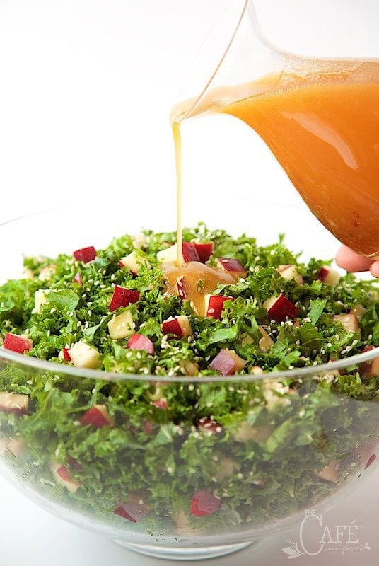 Vertical picture of Kale and Apple Salad in a glass bowl with Honey Ginger Dressing poured from a glass pitcher over the top.