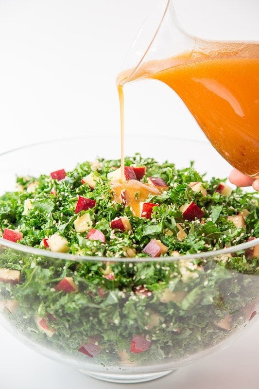 Vertical photo of a Kale and Apple Salad in a glass bowl with honey ginger dressing being poured on top from a glass carafe.